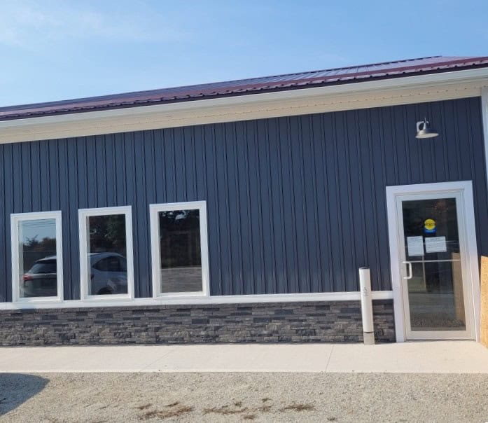 160821 A navy blue building with a dark stone trim. There is a walkway in front and gravel where people can park.
