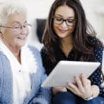Photo of older woman being shown by younger woman how to use a tablet.