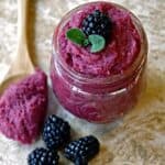 a purple homemade body scrub in a glass jar with three blackberries next to it