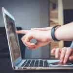 one hand pointing to the screen of a laptop and another hand pressing the navigation pad.