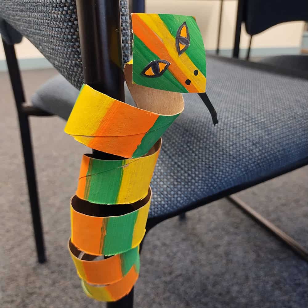 yellow, green and orange carboard snake wrapped around a chair leg.