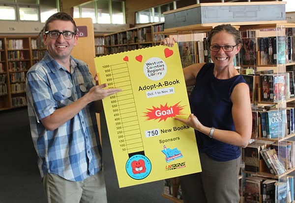 a man with glasses and a checked blue shirt and a woman with glasses, brown hair hold a yellow thermometer sign in a library.