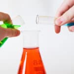 two hands pouring green and blue liquid from a beaker and a test tube into a larger beaker.