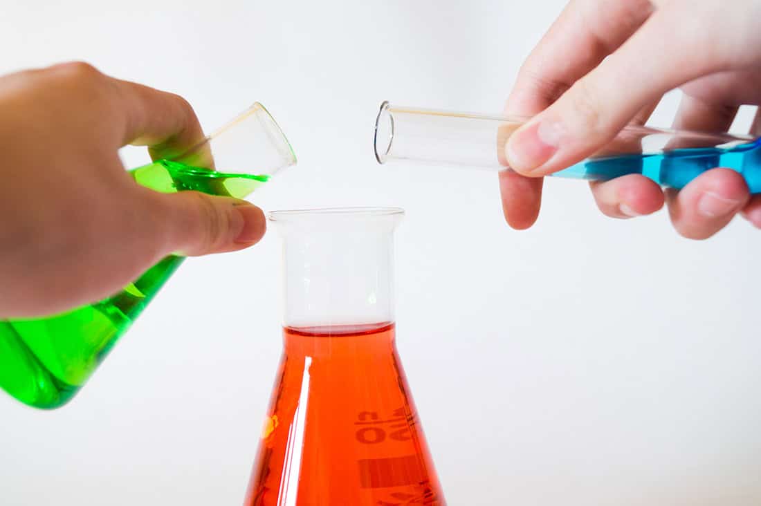 two hands pouring green and blue liquid from a beaker and a test tube into a larger beaker.