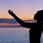 silhouette of a woman stretching out her arms as the sun sets over the ocean.