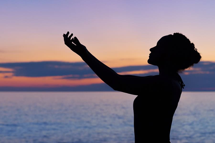 silhouette of a woman stretching out her arms as the sun sets over the ocean.