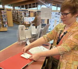 photo of a person with red hair, wearing glasses and a multi-coloured shirt over an orange T-shirt, placing a bookplate sticker on the inside page of a library book on a counter.