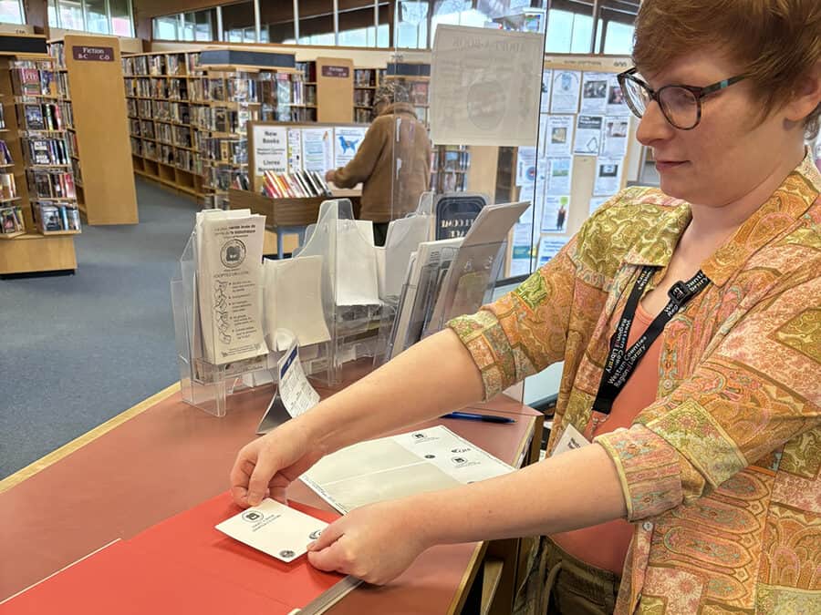 photo of a person with red hair, wearing glasses and a multi-coloured shirt over an orange T-shirt, placing a bookplate sticker on the inside page of a library book on a counter.