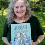 image of a woman with long, grey hair wearing a black top and holding a children's book with the text 'Josiah Wigglewart'