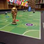 image of a mat soccer field with the World Cup trophy and round robots on it next to library shelves.