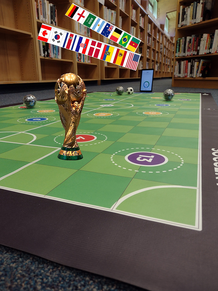 image of a mat soccer field with the World Cup trophy and round robots on it next to library shelves.