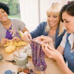 A group of three women knitting together. One woman has tan skin and dark curly hair, one has pale skin and long blonde hair, and the last has pale skin and wavy brown hair.