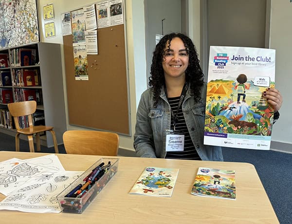 image of a young woman with dark curly hair wearing a jean jacket and striped shirt holds a Summer Reading Club poster while seated at a table