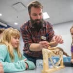 image of a man with a beard holding a roll of coins over a small wooden bridge with two girls watching.