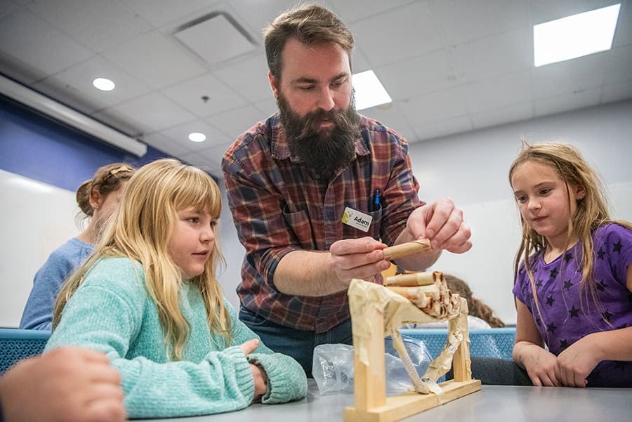 image of a man with a beard holding a roll of coins over a small wooden bridge with two girls watching.