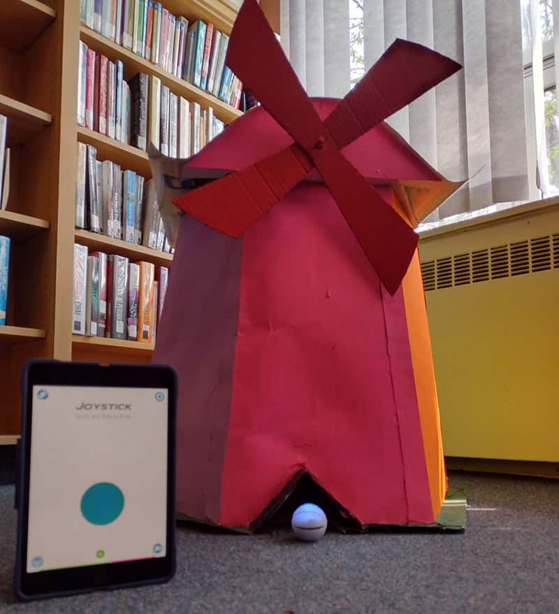 image of a red windmill on a carpeted floor in a library with a spherical robot and computer tablet in front of it.