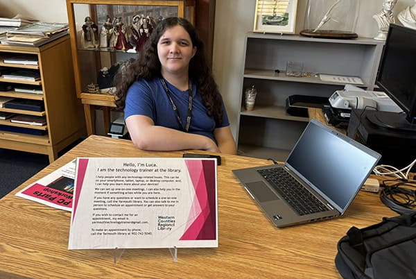 A woman sits at a desk with a sign and laptop computer on it.