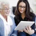 image of an older woman with white hair and glasses sitting next to a younger woman with dark hair and glasses holding a computer tablet.