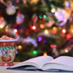 image of an open book on a table with e Christmas mug in front of a Christmas tree.