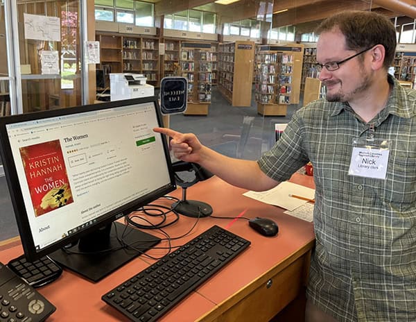 An image of a man in a checked, collared shirt with glasses and brown hair and a beard and mustache points to a computer screen at a desk.