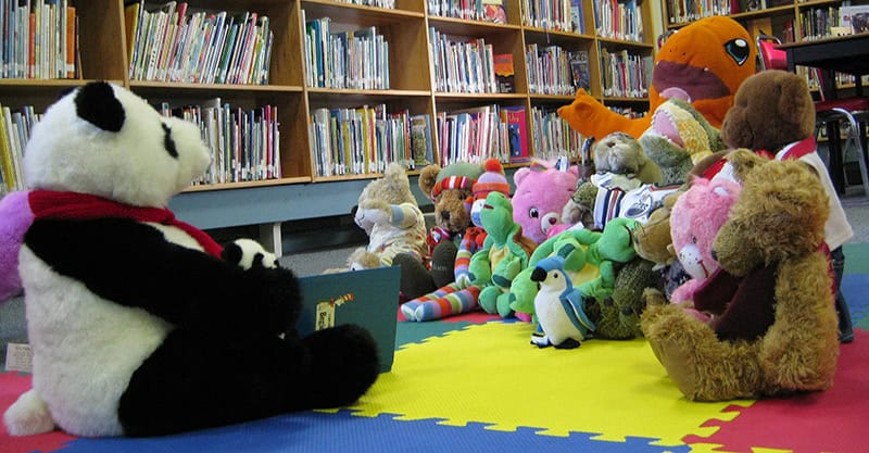 image of a stuffed panda bear holding a book in front of a bunch of stuffies on the library floor.