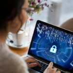 A young brunette woman with glasses typing on a laptop. The laptop screen has a lock on it to signify that her information is protected.