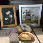 Needle felted frames pictures of daisies and a watering can full of flowers. On the table there are five needle felted coin purses.