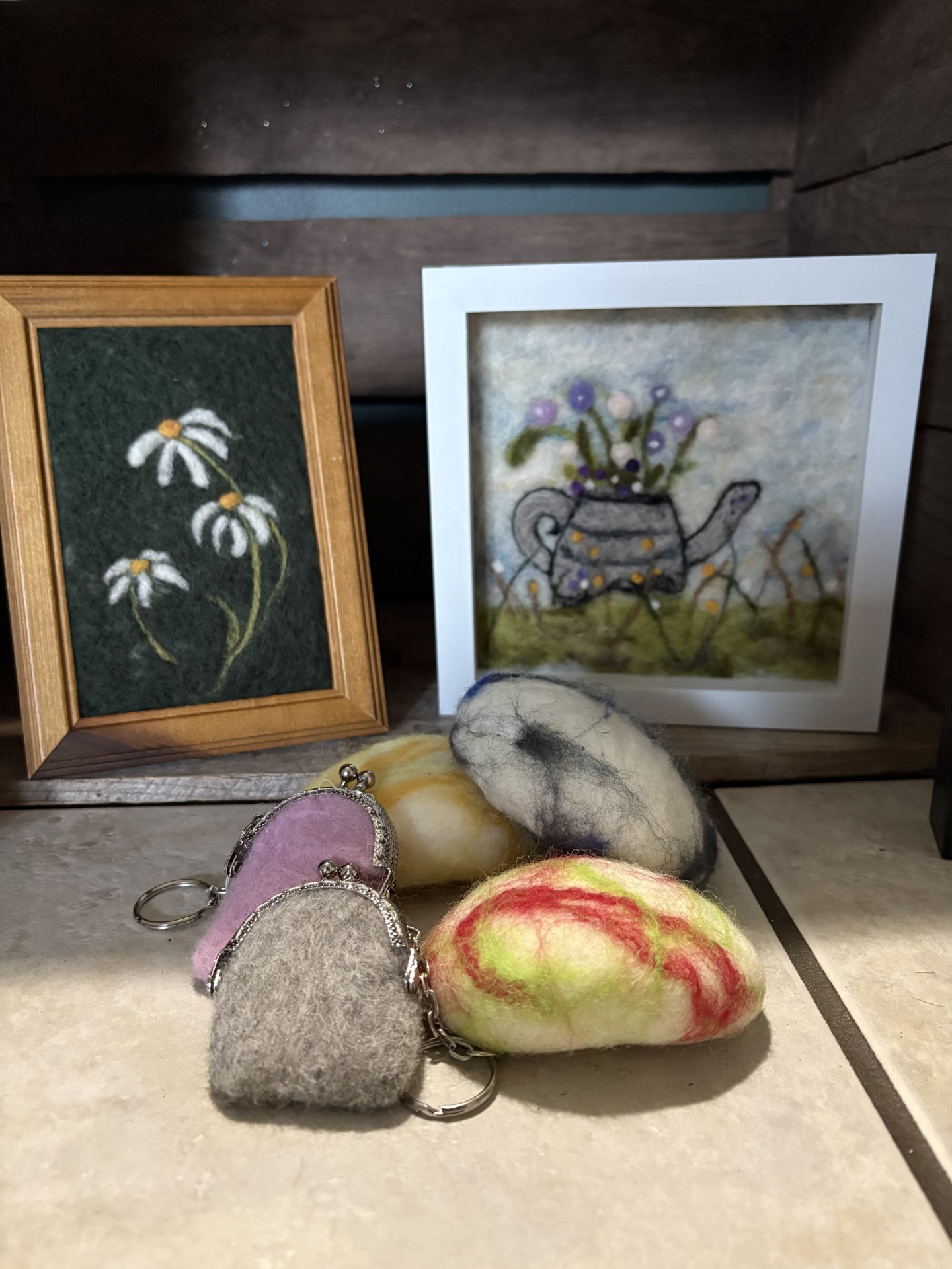 Needle felted frames pictures of daisies and a watering can full of flowers. On the table there are five needle felted coin purses.