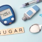 Items related to diabetes. Includes a blood sugar monitor, needle, finger prick, and a spoon of sugar. The background is light blue.