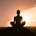 The silhouette of a woman sitting with her legs crossed, meditating on a grassy plain. Behind her is an orange sunset.