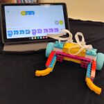 A plow made from LEGO Spike pieces. Behind it is a tablet with the LEGO Spike app and coding blocks displayed on it.