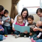 A group of toddlers of different gender and ethnicity sitting in a semi-circle. They are listening to a woman read a book. Some toddlers have parents with them.