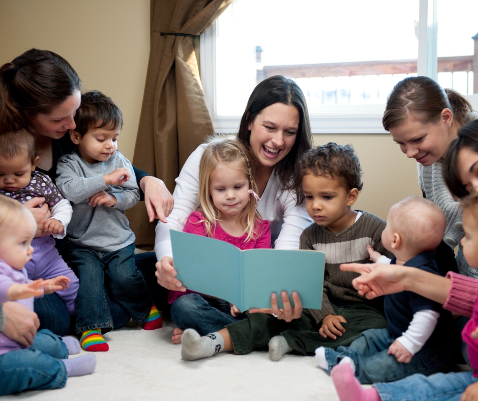 A group of toddlers of different gender and ethnicity sitting in a semi-circle. They are listening to a woman read a book. Some toddlers have parents with them.