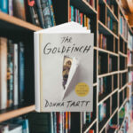 The cover is blank except for the middle where it looks like paper has been ripped away to reveal a painting of a goldfinch. Behind the cover are shelves of books.