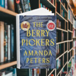 The cover has blueberry plant leaves in shades of blue. It also has two award stickers. Behind the cover are shelves of books.