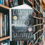 The cover is an illustration in blues and grays of a gloomy forest landscape. Behind the cover are shelves of books.