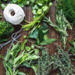 Various sprigs of herbs spread out on a wooden table. There is a spindle of white string and a pair of scissors to tie the herbs with.