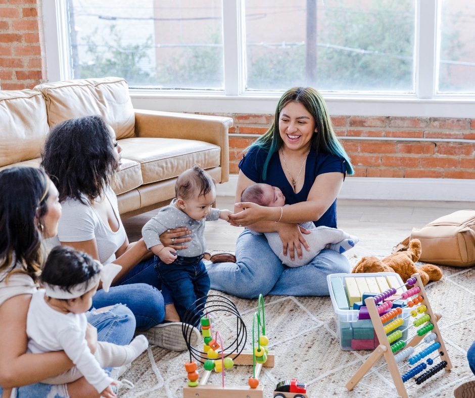 Three young women and their babies playing with block toys on the floor. They are smiling and talking to each other.