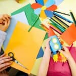 A table covered in construction paper, coloured pencils, and scissors. The hands of children are visible above the table. One child holds a paper rocket.