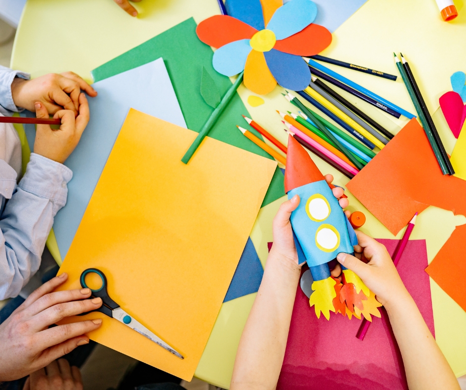 A table covered in construction paper, coloured pencils, and scissors. The hands of children are visible above the table. One child holds a paper rocket.
