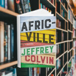 The cover of the book is made up of siding from different houses. Some are in good condition, some have chipping paint. Behind the cover are shelves of books.