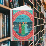 The cover features three cartoon women in regency period gowns. It is dark outside and there is a large manor behind them. Behind the cover are shelves of books.