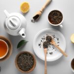 Various tea making objects spread out on a white table. There are containers of loose tea leaves, spoons of tea leaves, a teapot, and cups of steeped tea.
