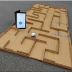 A maze made from cardboard. On top of it is a tablet with the Sphero app open and a Sphero robot.