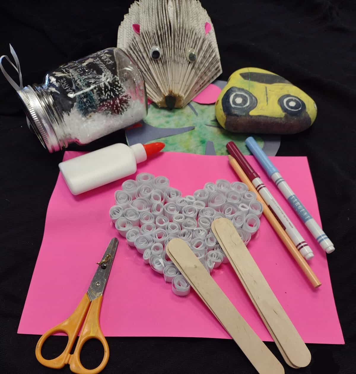 A display of many crafted items. There is a hedgehog made from book pages and a rock painted to look like a car. There are craft supplies scattered on the table.
