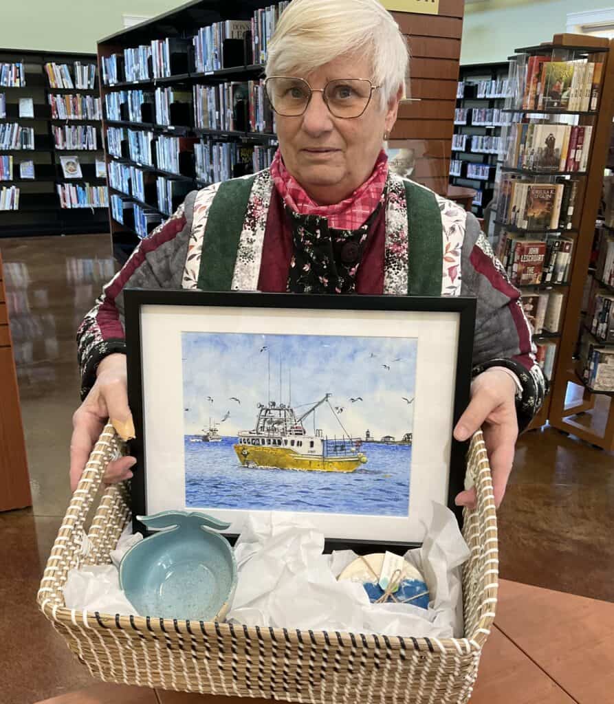 The winner of our adult Read Coast to Coast Challenge, Brenda F. She holds a wicker basket that has a painting of a ship, a whale ceramic bowl, and a set of coasters that look like waves.