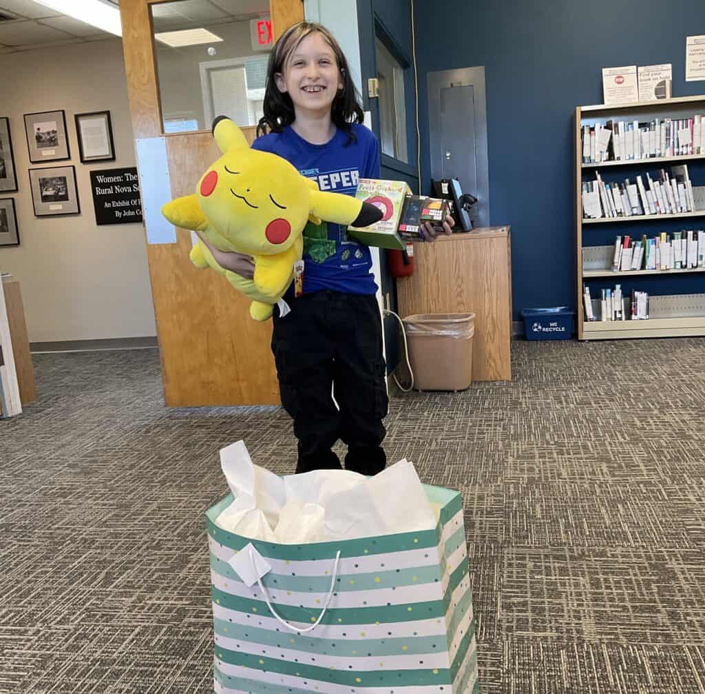 The youth winner of our Read Coast to Coast Challenge, Kit S. They are holding a Pikachu plushie, two Needoh squishies, and a Baby Three surprise box.