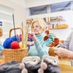 Three older white women sit at a table. They are admiring the socks they knitted.