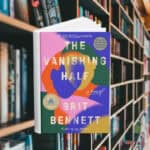 The cover features an abstract interpretation of two women leaning into each other. Their faces, bodies, and hair, are made of different coloured shapes. Behind the cover are shelves of books.