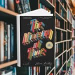 The cover features the title in 3d printed letters. The letters are all different colours. The background is black with small white dots that look like stars in space. Behind the cover are shelves of books.